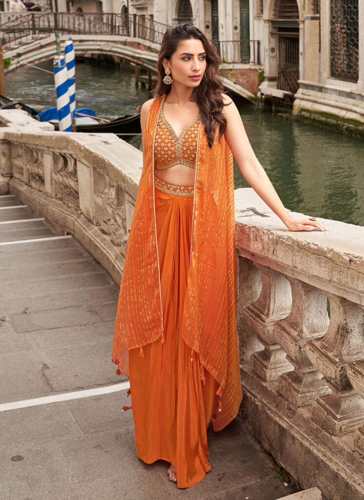 Indo Western Outfits In Chandni Chowk featuring an elegant orange drape-style outfit with an embroidered blouse and flowy cape, perfect for wedding functions, festive wear, and party wear at Ajmera Fashion Chandni Chowk.